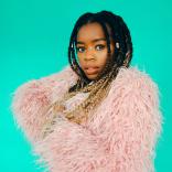 close up shot of a woman with long braided hair, styled with beads, posing against a teal background while wearing a fluffy pink coat.